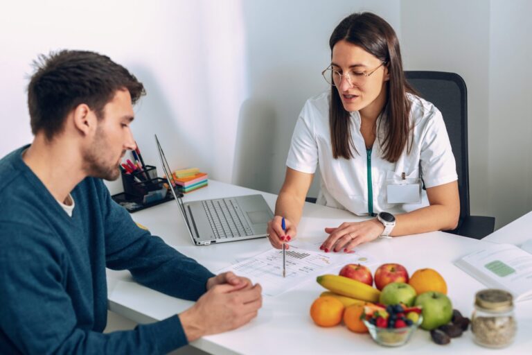 Ernährungsexperte sitzt mit Klienten am Tisch und berät ihn. Auf dem Tisch liegt gesundes Essen. medical,visiting,woman,young,caucasian,medic,dietitian,office,program,detox,professional,fit,loss,computer,routine,patient,indoors,diet,man,female,plan,table,profession,work,health,weight,control,medicine,calories,eating,people,food,lifestyle,doctor,help,nutrition,nutritionist,prescription,person,clinic,consultation,internet,sitting,care,women