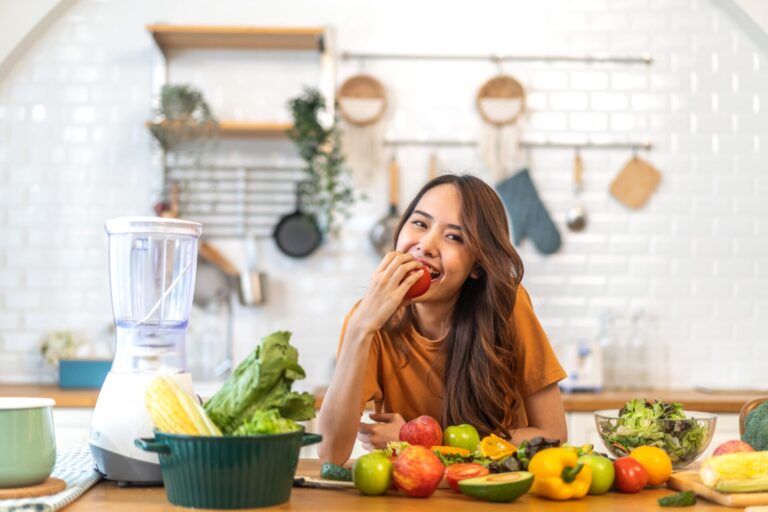 Frau sitzt an einem Tisch mit gesunden Nahrungsmitteln und beißt in einen Apfel. Sie sieht glücklich aus und lächelt. beauty,happy,dieting,hold,smile,dental,wellbeing,model,teeth,give,profile,cheerful,girl,delicious,dentist,vitamin,nutrition,present,care,strong,woman,fruit,skin,cute,summer,healthcare,apple,hungry,joy,vegetarian,eat,ripe,diet,organic,green,sexy,asian,health,vegan,clean,juice,portrait,food,face,mouth,healthy,juicy,fresh,sweet,stem