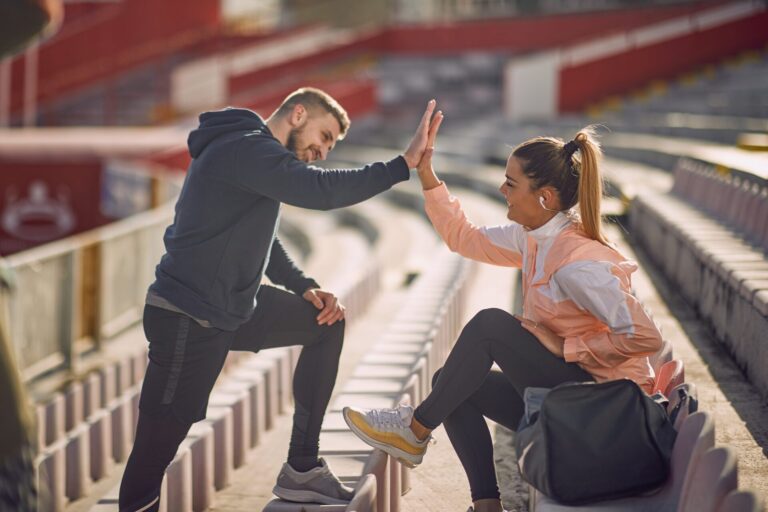 Ein Sportler und eine Sportlerin sitzen in einem Stadion und schlagen sich ab mit einem High Five. Beide lächeln, es könnte auch der Coach und eine Sportlerin sein. couple,woman,boyfriend,caucasian,happy,healthy life,slim,bag,togetherness,seats,professional,fit,relaxed,beautiful,high-five,outfit,joy,fitness,trainer,brunette,stadium,sweatshirt,man,female,relationship,flirting,smiling,attractive,looking each other,beard,tribune,satisfied,personal,sportswear,people,friends,lifestyle,girlfriend,standing,emotion,snickers,job,sport,male,sitting